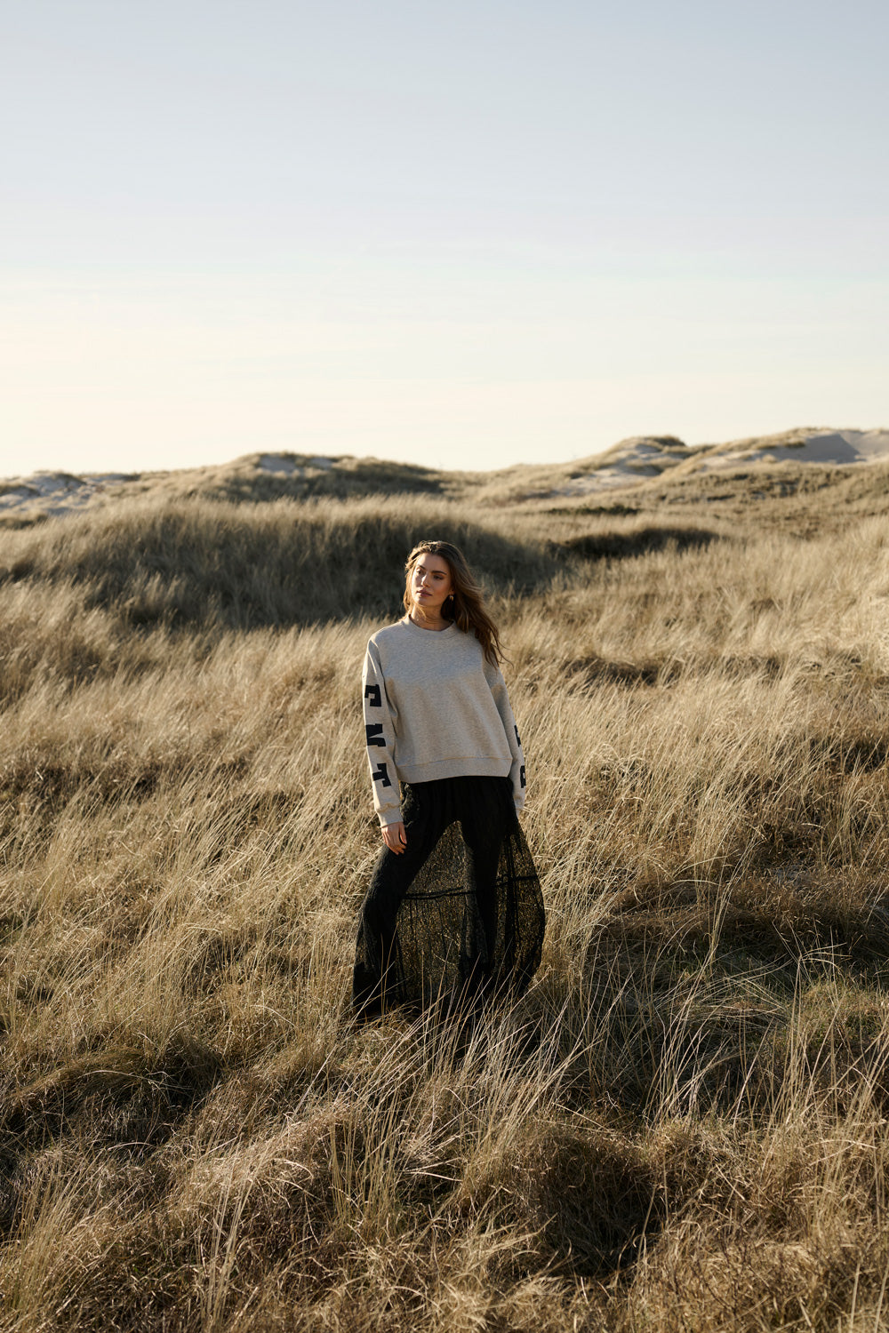 Eine Frau mit beigem Sweater und schwarzem langen Rock steht in einer Dünenlandschaft.