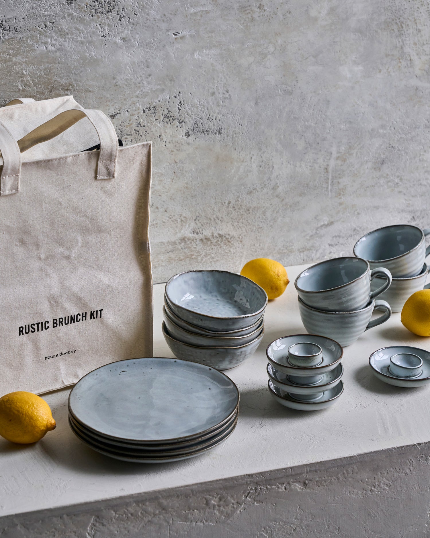 Blaugraues Geschirr steht gestapelt auf einem Tisch. Eine Leinentasche und Zitronen liegen daneben.