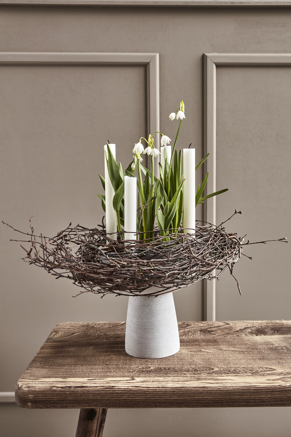 Ein Kranz aus Zweigen liegt auf einem Keramikkerzenständer. In der Mitte des Kranzes stehen Märzenbecher.